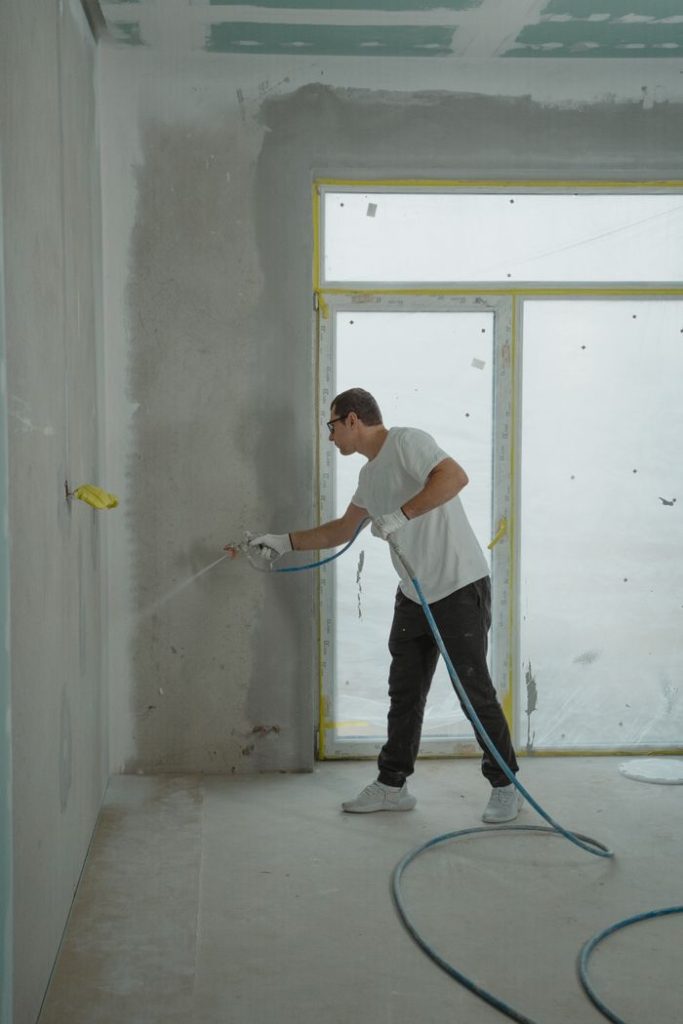 A man uses a spray gun to paint an interior wall in a room under renovation.