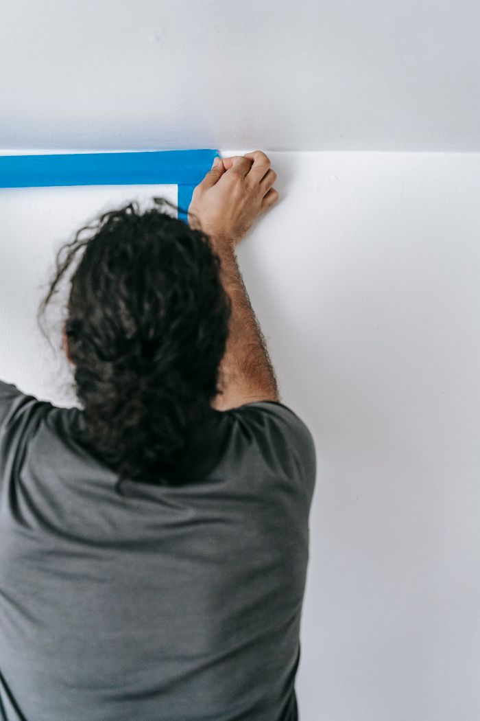 Back view of a man applying blue adhesive tape on a wall for a home painting project.